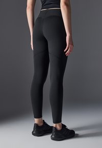 Leggings noirs avec un design ajusté, ornés de coutures et d'une taille haute, associés à des chaussures de sport noires.