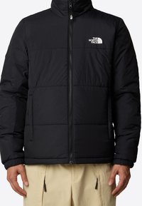 Chaqueta tipo puffer negra con cuello alzado, diseño acolchado, cremallera frontal y logo en el lado izquierdo del pecho. Cuenta con puños elásticos y bolsillos laterales.