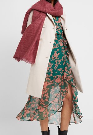 Femme portant un manteau beige boutonné sur une robe portefeuille fleurie verte, avec une grande écharpe rose poudré drapée autour des épaules.