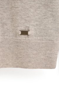 Beige Strickpullover mit einem gerippten Saum und einem kleinen Metall-Logo-Anhänger mit der Aufschrift "POLO CLUB." Weiche Textur und leichtes Design.