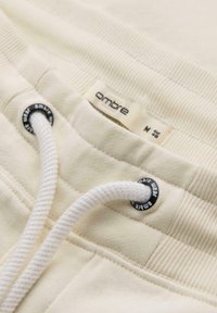Gros plan sur la ceinture de pantalon de survêtement beige avec un cordon blanc passé à travers des œillets métalliques marqués et une étiquette de taille indiquant "ombre M".
