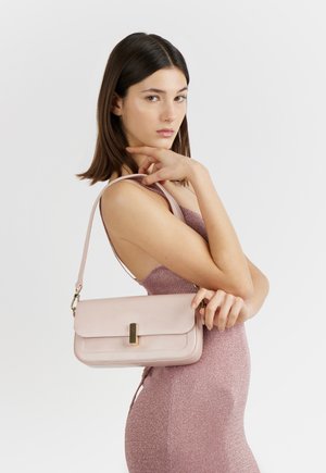 Giovane donna in abito rosa senza maniche tiene una borsa a tracolla rosa chiaro con fibbia dorata su uno sfondo bianco uniforme.