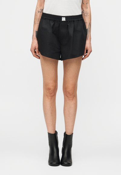 Personne portant un short à taille élastique noir, des bottines noires et un haut blanc, debout devant un fond gris clair uni.