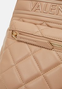 Beige quilted leather bag with a textured surface, featuring a gold zippered pocket and embossed logo. Smooth finish, minimalist design.