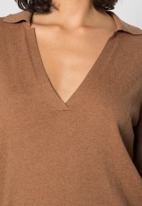 Kobieta w brązowym swetrze z dekoltem w serek i kołnierzykiem, odsłaniająca dekolt i górną część klatki piersiowej.