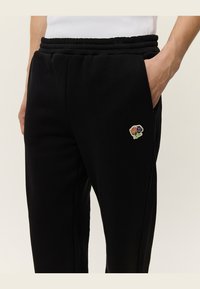 Pantalones de chándal negros con cintura elástica y bolsillos laterales, que presentan un pequeño parche bordado de una rosa en colores púrpura, naranja y verde.