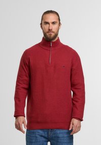 Roter, strukturierter Pullover mit hohem Kragen und halbem Reißverschluss, mit einem kleinen Logo auf der Brust. Getragen mit blauen Jeans.