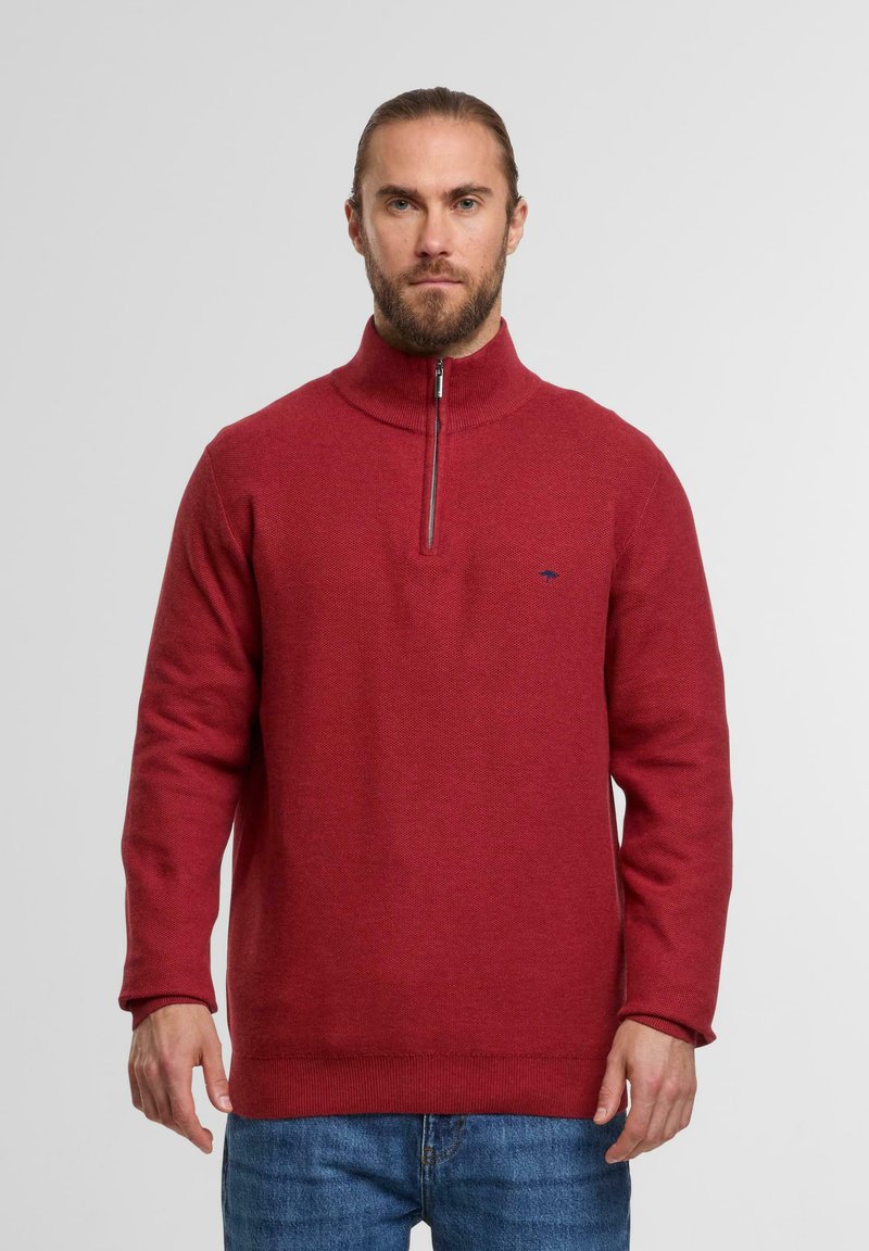 Roter, strukturierter Pullover mit hohem Kragen und halbem Reißverschluss, mit einem kleinen Logo auf der Brust. Getragen mit blauen Jeans.