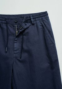 Pantalones azul marino con cintura elástica, botón, bragueta con cremallera parcialmente abierta, cordones y bolsillos laterales sobre fondo claro.