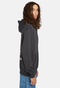 La sudadera gris cuenta con un bolsillo frontal, capucha con cordón y puños acanalados. Se encuentra junto a unos jeans azul claro.