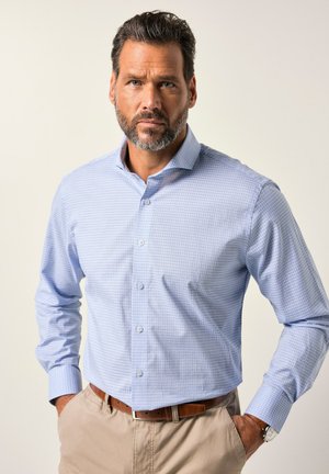 Ein mittelalter Mann mit Bart, der ein hellblaues karriertes Hemd, beige Hosen, einen braunen Gürtel und eine Armbanduhr trägt, steht mit den Händen in den Taschen.