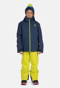 Giacca impermeabile blu navy con cerniere giallo neon e pantaloni da neve; presenta un cappuccio aderente e dettagli neri. Berretto lavorato a maglia con pompon luminoso in cima.