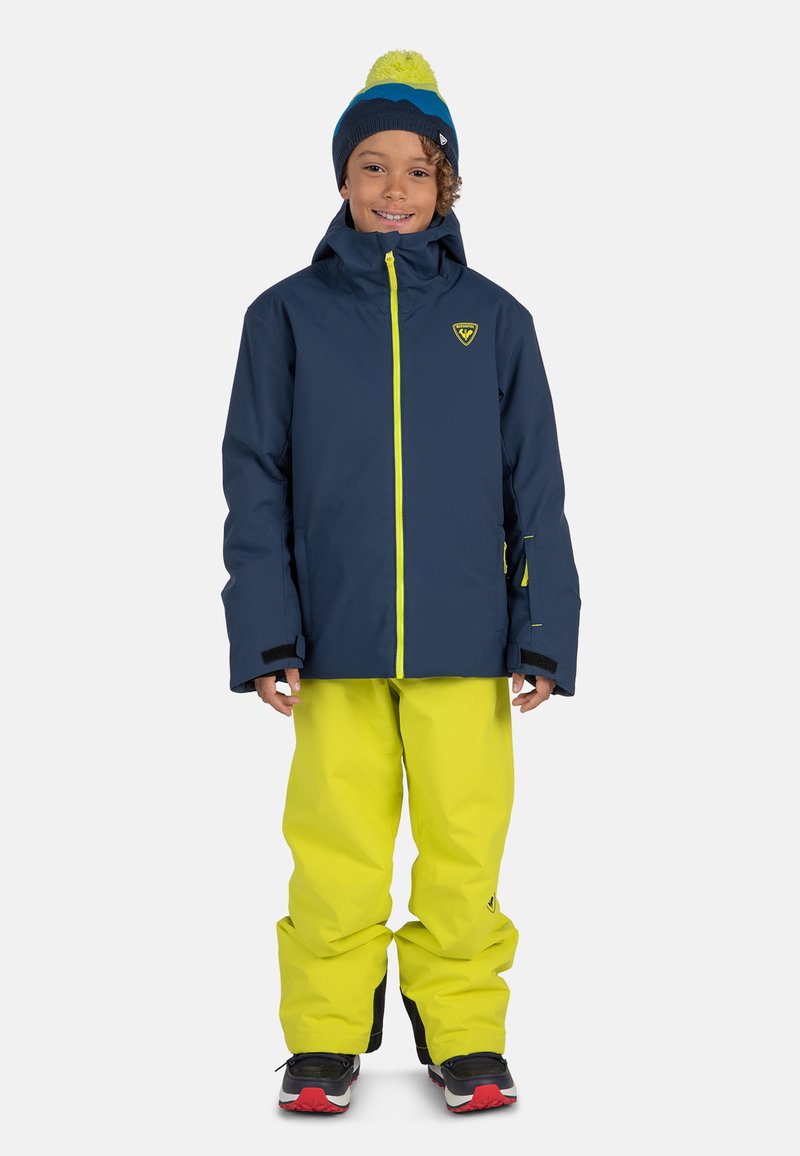 Giacca impermeabile blu navy con cerniere giallo neon e pantaloni da neve; presenta un cappuccio aderente e dettagli neri. Berretto lavorato a maglia con pompon luminoso in cima.