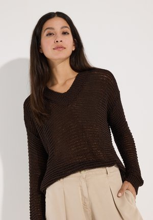 Vrouw met lang donker haar die een losse donkerbruine gebreide trui en lichtbeige broek draagt, met één hand in de zak.