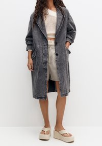 Harmaa farkkukangasta pitkä takki, jossa on lovellinen kaulus, kaksi etutaskua ja kolme nappia. Päälle puettuna vohvelikuvioinen kerma-pitkä hiha crop top ja vaaleat sortsit.