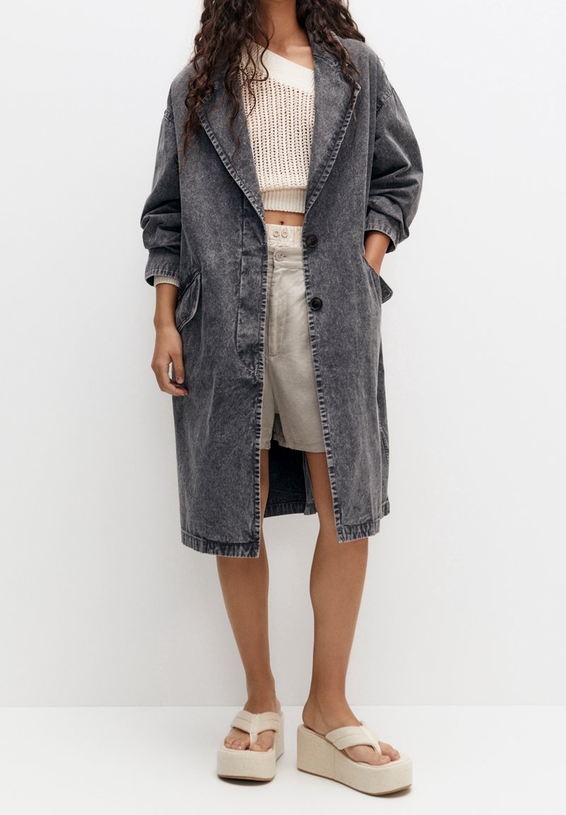 Harmaa farkkukangasta pitkä takki, jossa on lovellinen kaulus, kaksi etutaskua ja kolme nappia. Päälle puettuna vohvelikuvioinen kerma-pitkä hiha crop top ja vaaleat sortsit.