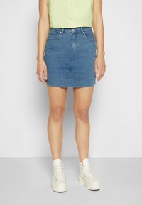 Saia mini de denim com bainha desfiada, apresentando uma cor azul clara, design ajustado e detalhes visíveis nos bolsos frontais. Combinada com tênis brancos de cano alto.