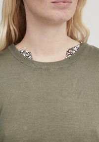 Oliven grøn strikket sweater med rund halsudskæring. Et blomstermønstret stof titter frem fra under kraven og tilføjer kontrast.