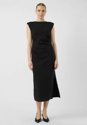 Abito nero aderente con maniche a cappuccio, asimmetrico lungo il ginocchio sul lato sinistro, texture liscia, design drappeggiato in vita, abbinato a scarpe basse nere.