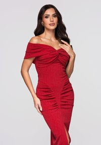 Czerwona, dopasowana sukienka z opuszczanymi ramionami, z marszczonym wzorem i rozcięciem do uda. Materiał ma błyszczącą fakturę. Widoczne są złote dodatki.