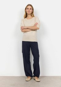 Haut beige tricoté à manches courtes, porté avec un pantalon cargo bleu marine doté de poches latérales. Des chaussures plates de couleur claire complètent la tenue.