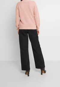 Pull rose clair en tricot avec un motif texturé, associé à un pantalon large noir et des chaussures à talons hauts foncées. Vue de dos.