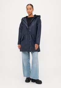 Temno modra vodoodporna jakna s kapuco, z sprednjimi gumbi na pritiskač, dvema stranskima žepekoma in obloženim ovratnikom, v kombinaciji z svetlo modrimi širokimi jeans hlačami.