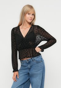 Zwarte sheer blouse met stippen, met een wikkelontwerp, lange mouwen en een peplumrand, gecombineerd met lichtblauwe high-waisted jeans.