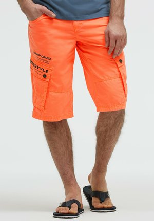 Mann trägt leuchtend orangefarbene Cargo-Shorts mit gedrucktem Text auf der linken Tasche und schwarze Flip-Flops und steht vor einem schlichten Hintergrund.