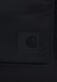 Geantă din material textil negru cu o suprafață texturată. Prezintă un patch pătrat, ridicat, cu un logo de culoare gri închis. Linii curate și design minimalistic.
