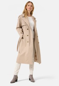 Trench coat beige con design doppiopetto, bottoni dorati e tasche laterali. Tessuto morbido, lunghezza al ginocchio, abbinato a pantaloni fantasia e stivaletti grigi alla caviglia.