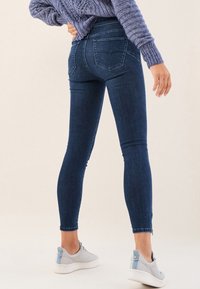 Jeans ajustados de color azul oscuro con cintura alta, diseño de cinco bolsillos y detalles de costura sutiles. Combinados con zapatillas de deporte gris claro.