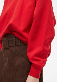 Röd sweatshirt med avslappnad passform och långa ärmar, matchad med bruna mockabyxor med hög midja som har en bälte och sydda detaljer.