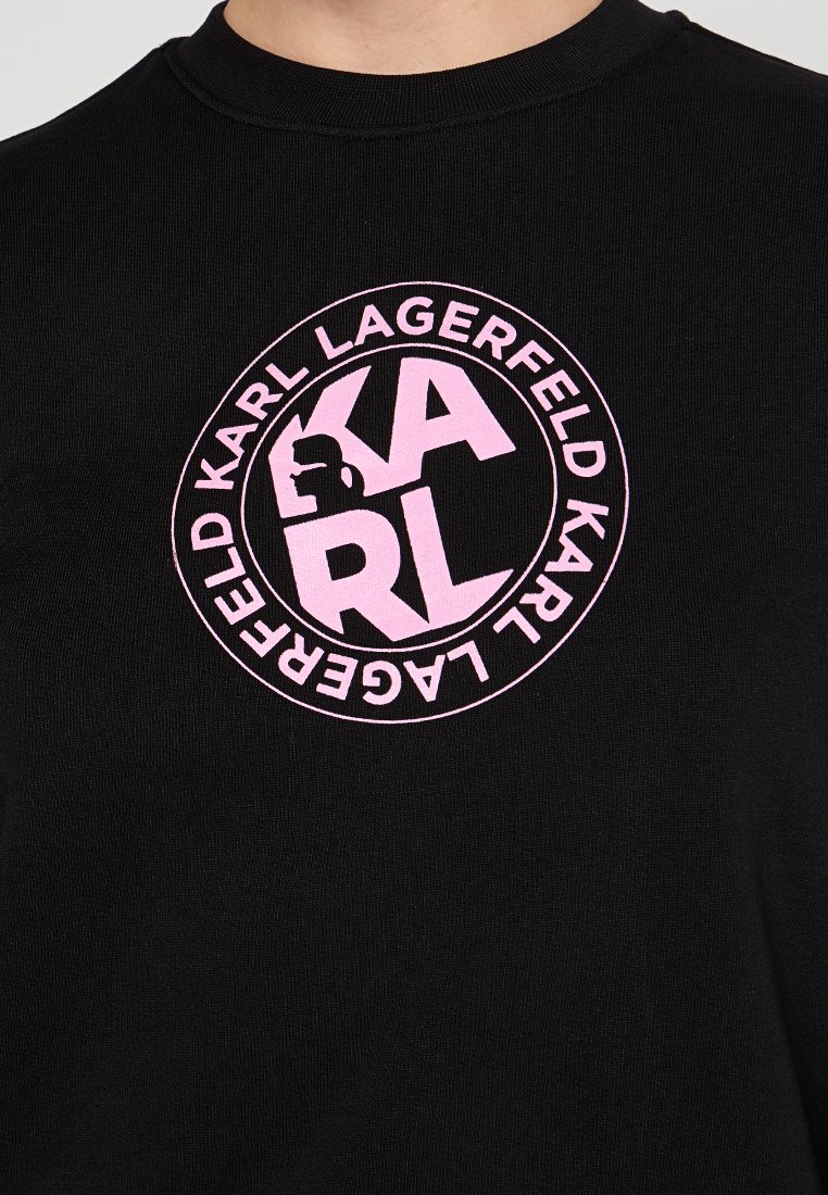 Camiseta de algodón negra con un gráfico circular en rosa que incluye el texto "KARL LAGERFELD" y una silueta, creando un contraste audaz.