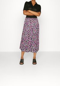 Femme debout, bras croisés, portant un top noir à manches courtes, une jupe midi noire à fleurs et des sandales noires ouvertes à l27avant.