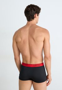 Zwarte boxer shorts met een rode Calvin Klein tailleband, met een aansluitende pasvorm en een gladde textuur, weergegeven vanuit een achteraanzicht.