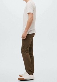Světlé béžové tričko s krátkým rukávem, hnědé slim-fit kalhoty a bílé tenisky s hnědými gumovými podrážkami stojí na neutrálním pozadí.
