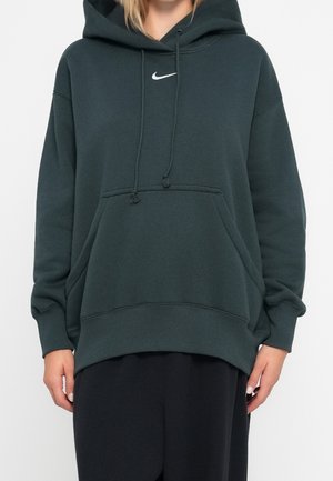 Osoba nosząca ciemnozieloną bluzę z kapturem Nike z przednią kieszenią i sznurkami, w połączeniu z czarnymi spodniami, widoczna od ramion do bioder.