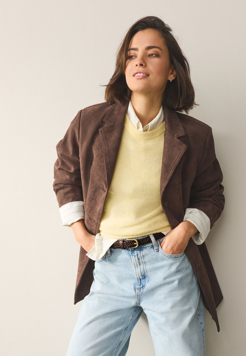 Jeune femme aux cheveux courts et foncés portant un blazer marron, un pull jaune clair, une chemise blanche et un jean bleu clair, debout les mains dans les poches.