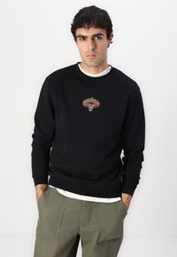 Black sweatshirt with an embroidered graphic featuring a skull and sombrero. Crew neckline, long sleeves, and ribbed cuffs. Worn over a white shirt.