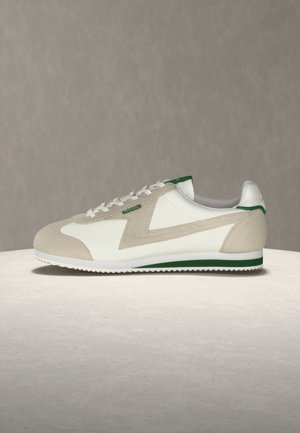 Hvite og beige lave sneakers med grønne detaljer, semskede overlegg, snøring og såle med sikksakkmønster, står på en lys overflate.