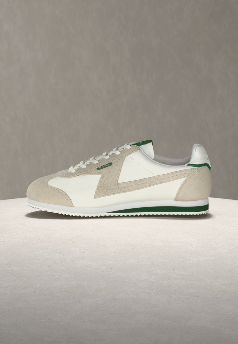 Baskets basses blanches et beige avec accents verts, superpositions en daim, fermeture à lacets et semelle en caoutchouc en zigzag, posées sur une surface claire.