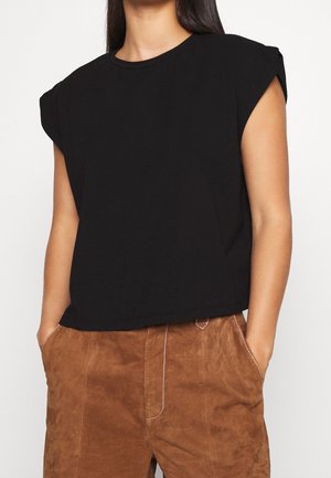 Vrouw met een mouwloze zwarte top en bruine suèdé broek met handen in de zakken, gefotografeerd van schouders tot halverwege de dij.