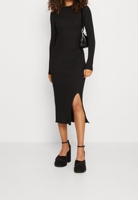 Schwarzes geripptes Midikleid mit langen Ärmeln, seitlichem Schlitz und tailliertem Schnitt. Kombiniert mit schwarzen Plateauschuhen und einer kleinen schwarzen Handtasche.