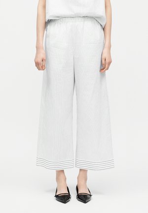 Žena v bílých širokých 3/4 kalhotách s tenkými vertikálními pruhy a černých lodičkách s otevřenou patou a špičatou špičkou.