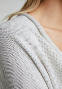 Pull en tricot blanc avec un design ample, une texture douce et un léger éclat, doté d'un col en V et d'un décolleté drapé.