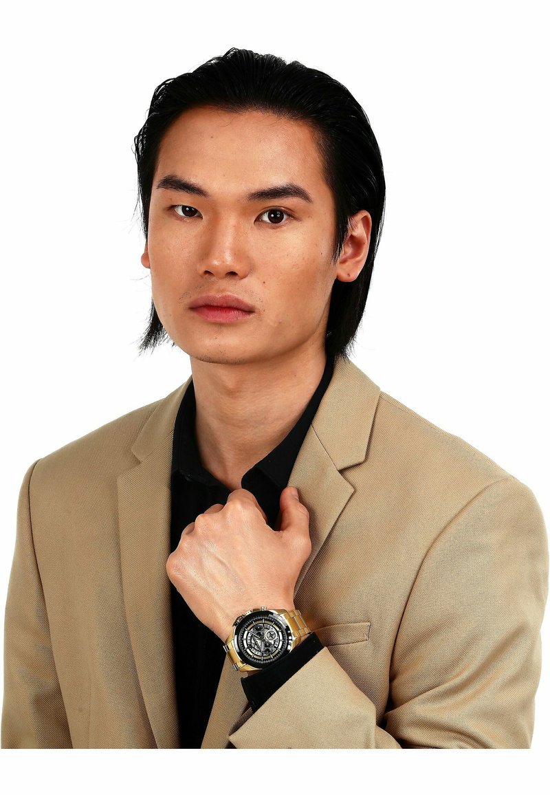 Gold- und Silber-Analog-Uhr mit schwarzem Zifferblatt und filigranen Details. Am Handgelenk getragen, mit einem Metallarmband und strukturiertem Gehäuse.