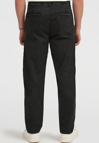 Mand iført sorte lige ben bukser med baglommer og hvide sneakers, vist fra baggrunden mod en ensfarvet baggrund.