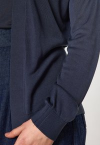 Cardigan bleu marine avec des poignets et un ourlet côtelés, fabriqué à partir d'un tissu tricoté texturé, présentant une ouverture devant et une coupe décontractée.