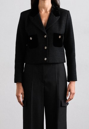 Frau trägt einen taillierten schwarzen Blazer mit goldenen Knöpfen und schwarze, weit geschnittene Hosen mit Klappentaschen, steht vor einem einfarbigen Hintergrund.